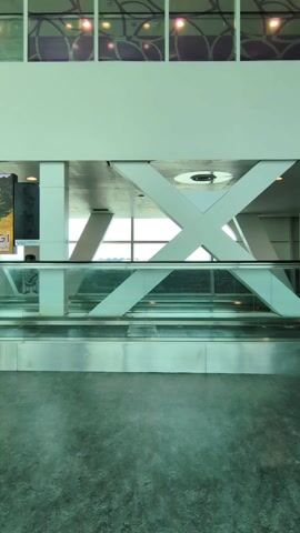 The video shows a moving walkway in what appears to be an airport terminal or a similar large public space. Several people walk across the moving walkway in both directions. The architectural features of the space include large structural beams and a decorative window pattern above. The floor is made of a dark, possibly polished material that reflects the light. A poster can be seen on the left side of the video. The scene is brightly lit, and the overall color palette is dominated by white, silver, and light green.