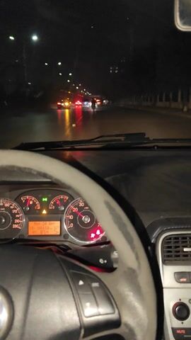 A video shows the interior of a car driving at night. The dashboard is visible, including the speedometer, fuel gauge, and digital display. A hand appears briefly in the frame, holding up three fingers. The steering wheel is gray and worn. The video then cuts to a shot of a person in the back seat, leaning on the car door.