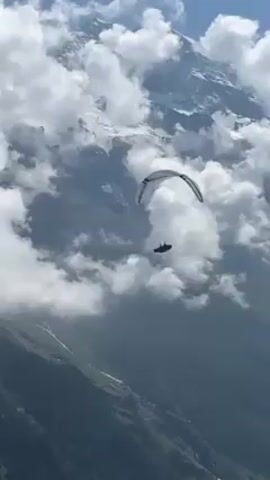 A paraglider is flying through a mountainous region with heavy cloud cover. The paraglider is white with blue accents. The background consists of mountain peaks covered in snow and lush green hillsides. The clouds are white and billowy, partially obscuring the mountain views.