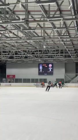 The video shows an indoor ice hockey rink during a game. A scoreboard hangs above the ice displaying the score as 2 to 3, with the clock showing 00:00. Several players in green and orange jerseys are on the ice, some engaged in play near the goal. The rink is illuminated by overhead lights. The overall environment appears to be a sports facility with metal scaffolding and ceiling structures visible.