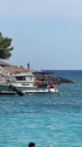 A boat is docked near a rocky shore with a person standing on the rocks. The water is blue and clear, with buoys marking an area in the foreground. The sky is clear and blue.