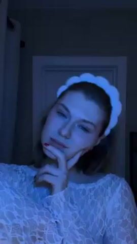 A woman is shown in a medium close-up shot. She is wearing a white lace shirt and a white headband. The background is a dark room with a closed door and a curtain. The lighting is dark, with a blue hue. The woman moves her hands and speaks to the camera.