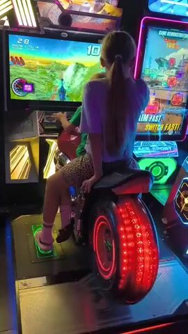 The video shows a girl and a younger child sitting on a motorcycle arcade game. The girl is in the back, and the child is in front, holding onto the handlebars. They are both facing the screen of the game, which displays a racing game with graphics of a road and mountains. The motorcycle is red and black, with glowing red lights on the back tire. The arcade machine is brightly lit with neon lights. The girl is wearing a purple t-shirt and patterned shorts. The child is wearing a green t-shirt and patterned shorts. The video is shot from a side angle, focusing on the children and the game.