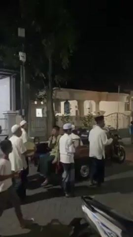 Here is a detailed description of the video:

The video shows a procession of people, predominantly men and boys, moving down a street at night. The group appears to be engaged in some kind of cultural or religious event. Many of the men are dressed in traditional Muslim attire, including white robes and head coverings, while some of the boys are wearing more casual clothing. A man is seated in the bed of a small truck and is singing into a microphone.

Several people are carrying light sticks. The environment is a residential street with houses and trees visible in the background. The lighting is provided by streetlights and the illumination from the houses, making the scene relatively well-lit despite the darkness.