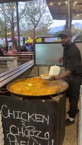 Here's a description of the video:

A man is standing beside a large paella pan, serving food into a takeout container. He is smiling. He is wearing a black chef's coat, a cap, and white sneakers. The paella is in a large, flat, round pan. The paella contains rice, green peas, tomatoes, and lemon wedges. Steam is rising from the paella. A black chalkboard in front of the pan says "CHICKEN & CHORIZO". In the background, there are picnic tables with people seated. There is an awning overhead with string lights. Trees are visible in the background. A mountain range is visible in the far background.