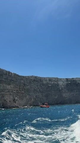 The video shows a view from a boat moving on the sea toward a cliff. The sea is a deep blue, with white foam forming behind the boat. The cliff is tall and grey, with a rugged, layered appearance. Several boats are docked near the base of the cliff, including an orange one. The sky is clear and blue.