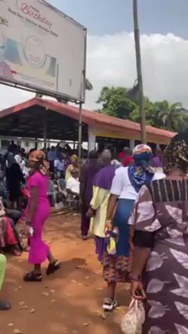 The video depicts a crowded outdoor event, possibly a celebration or ceremony, under a partially covered area. People of various ages and genders are walking and standing around. Many individuals are dressed in colorful and patterned clothing, with some wearing traditional attire and headwraps. There's a large sign that says "Happy Birthday Most Rev. Valerian Maduka Okeke." In the background, there's a tall building with multiple windows and trees. The ground appears to be a dirt or clay surface. The atmosphere seems lively, with people interacting and moving about.