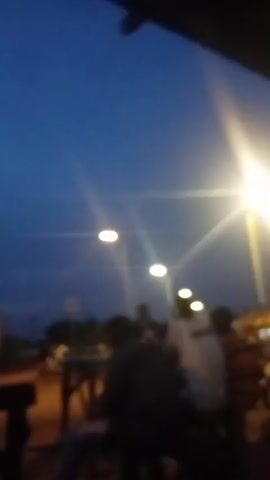 The video shows a nighttime outdoor scene with a bright light pole at the center, creating a starburst effect. Several people are visible, some sitting and others standing, in the lower portion of the frame. The environment appears to be a public area with some structures or tents in the background. The sky is dark blue, indicating it is either dusk or night. The video is shaky, suggesting it was handheld.