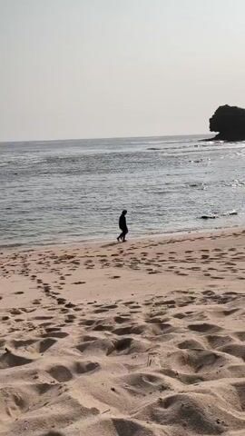 The video shows a sandy beach with footprints in the sand. The ocean is visible in the background, with waves rolling towards the shore. There is a large rock formation in the distance. Two people are walking along the water's edge. One person is walking slightly ahead of the other, and they appear to be interacting. The sun is low in the sky, casting a warm glow over the scene.