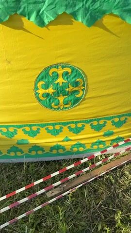 The video shows a close-up view of a yellow yurt decorated with green trim and patterns. The yurt has a circular emblem with green patterns in the center. There are two flags flying from the top of the yurt: a Russian flag and an Uzbek flag. The video also shows grass and some wooden planks on the ground. The flags appear to be blowing in the wind. The video is shot from a low angle, looking up at the yurt.
