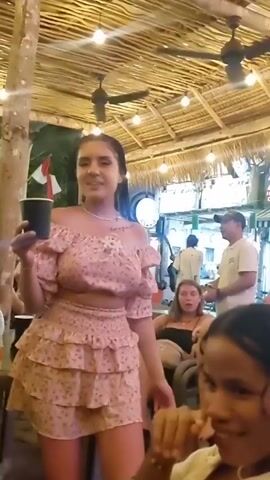 The video is taken from a low angle, looking up at a group of people sitting at tables under a thatched roof. A woman in a pink floral off-the-shoulder crop top and matching skirt is holding a black cup. She raises her cup and yells "Hi!" into the camera. The hand of the person filming is in the foreground, partly obstructing the view. Other people are seated around the table, and there are people moving around in the background. The environment appears to be an outdoor bar or restaurant, with lights strung across the thatched roof and trees nearby.