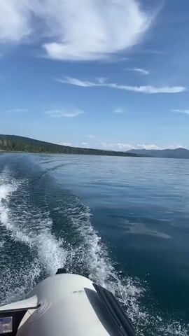The video shows a view from a boat moving through the water. The camera is pointed forward and slightly to the left, capturing the wake of the boat. The water is a deep blue-green and calm, with small ripples. In the distance, a treeline stretches across the horizon, followed by rolling hills or mountains under a bright blue sky with scattered white clouds. The front of a white boat is visible in the lower right corner of the frame. The video appears to be a short clip, possibly a screen recording, as indicated by the timestamp at the beginning.