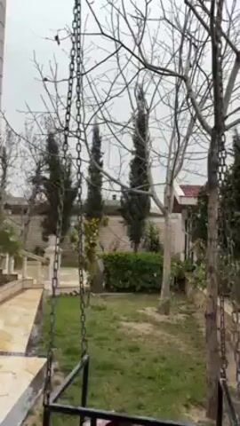 The video shows a backyard scene. There is a swing hanging from a leafless tree. The swing appears to have a white cushion and a maroon blanket on it. The backyard has a stone pathway, grass, various trees, bushes and plants. A building with light-colored walls can be seen in the background. The sky is overcast. The camera pans slowly across the scene.