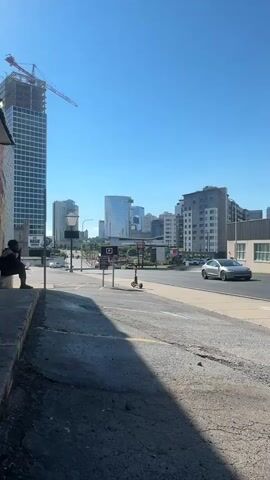 The video shows a street scene in a city. In the foreground on the left, a person is sitting on a ledge. To the right of the person, there is a street with a cracked asphalt surface. Further down the street, there are signs, including a parking sign and a sign for "USED CARS". A red electric scooter is parked near the parking sign. In the background, there are several modern buildings, including a tall skyscraper with a crane on top, indicating ongoing construction. The sky is clear and blue. Cars are visible on the street in the middle ground. The lighting suggests it is daytime.