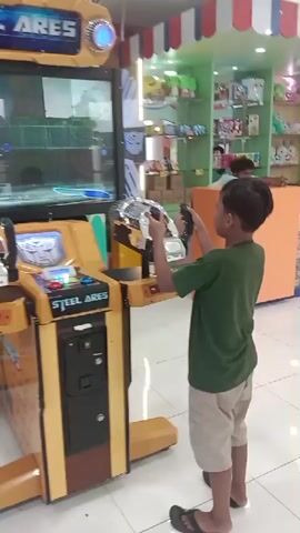 A young boy is playing an arcade game called "Steel Ares" in what appears to be an indoor entertainment area. The game machine is predominantly yellow with a large screen displaying racing game footage. The boy, wearing a green shirt and beige shorts, is holding the game's steering wheel and enthusiastically engaging with the game. The surrounding environment features a brightly lit interior with tiled floors and displays of various items and toys on shelves in the background, indicating a possible retail or entertainment space.