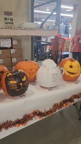 The video shows a long white table filled with creatively decorated pumpkins, numbered for some kind of contest. The table is decorated with orange and black tinsel. The pumpkins include a black pumpkin with a cat face, a white pumpkin painted like a pumpkin spice latte, and a Frankenstein pumpkin painted green. Behind the table are shelves stacked with boxes and other items, suggesting this takes place in a storage area or warehouse.  A few people are visible in the background.