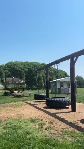 The video shows a tire swing set on a sunny day. The swing set is made of two wooden posts supporting a wooden beam from which three large tractor tires are suspended by chains. In the background, there is a small building with a mural painted on it and some trees. There are also some old farm equipment in the distance. The ground is covered with grass and dirt. The sky is clear blue.