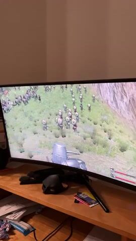 This is a video of a computer monitor displaying a video game. The game appears to be a real-time strategy or action game set in a medieval or fantasy world. A player character, a knight on horseback, is shown in the foreground, wielding a lance. In the background, a large army of soldiers, also appearing to be on horseback, is engaged in battle on a grassy hillside. The camera angle is from a third-person perspective, following the player character. The monitor is curved, suggesting it is a widescreen display. On the desk in front of the monitor, there are several items, including a black mask, pens, and some papers. The lighting in the room is dim, with the light from the monitor being the primary light source.