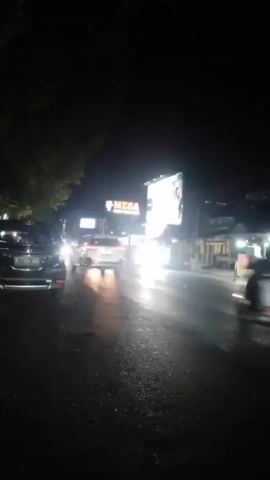 The video shows a nighttime street scene. Traffic is moving on a wet road, reflecting the lights of vehicles and a large illuminated billboard advertising "MEGA". Several cars and motorcycles are visible. One car in the foreground appears to be a dark SUV. A motorcycle with a rider passes by. The surrounding area includes what appears to be roadside stalls or shops with lights on. The overall atmosphere is that of a busy street at night.