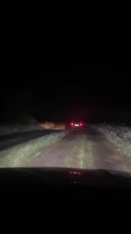 The video shows a first-person perspective from inside a vehicle at night. The headlights of the vehicle illuminate a dirt road. In the distance, another vehicle with its red taillights illuminated is visible. The road is surrounded by dark vegetation. The vehicle appears to be moving forward, following the lead vehicle.