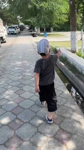 A young boy wearing a grey t-shirt, black shorts, a grey baseball cap, and sandals walks down a paved sidewalk. The sidewalk is lined with a concrete irrigation ditch on his right and trees and a street with parked cars on his left. He is walking away from the camera.