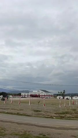 The video pans across an open, undeveloped field under a cloudy sky. In the distance, there is a large, modern building with a white facade and a red roof, possibly a government building or a public facility. In front of the building, several Indonesian flags are displayed on poles. Scattered across the field are smaller, white buildings, also possibly part of a larger complex. The foreground shows a dirt road and some sparse vegetation. Power lines are visible strung across the scene. The overall impression is one of a developing or underutilized area.