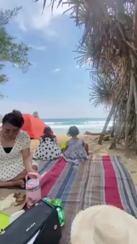 A family or friend group is having a picnic on a beach. There are two small children sitting on a blanket, looking out at the ocean. A woman in a white and black polka dot dress is preparing food on the blanket, while another woman in a floral shirt sits nearby eating. There are several containers of food, bags, and other items spread out on the blanket. Behind them, there are several tents set up on the beach, and people can be seen relaxing on lounge chairs. The weather is sunny and the sky is blue. The ocean has gentle waves.