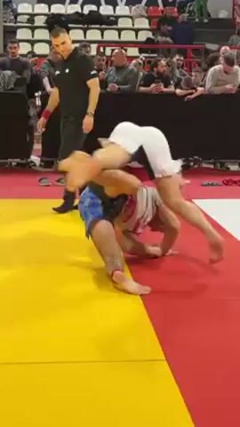 Two men are grappling on a yellow and red mat. One man wears a red shirt, white shorts, and has a beard. The other man wears a white rash guard and shorts with blue and white designs. The man in the red shirt is on top, holding the other man in a hold. A referee stands nearby, watching the match. Behind them, a crowd of people is watching from the stands. There are trophies on a table in the background.