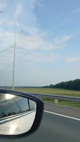 The video, recorded from the interior of a moving vehicle, shows a side-view mirror reflecting a bright sky with scattered clouds and the sun. Beyond the mirror, a green field stretches into the distance, bordered by a line of trees. A metal guardrail runs along the side of a road, indicating the vehicle is traveling on a highway. The camera pans slightly, capturing the passing landscape under a clear blue sky.