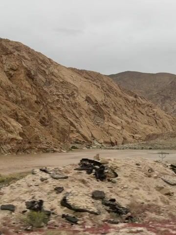 A moving shot captures a desolate, rocky landscape under an overcast sky. The camera pans across a wide, muddy riverbed bordered by towering, jagged, reddish-brown mountains. Sparse green vegetation and scattered rocks dot the foreground and the riverbanks. The scene is devoid of human presence. The overall impression is one of a rugged, untouched natural environment.