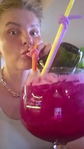 A woman is shown with a large purple drink, possibly a cocktail, in a goblet. She is holding two straws, one orange and one yellow, and appears to be drinking from the orange straw. The woman has blond hair styled up, and is wearing a necklace and earrings. She is looking at the camera with a surprised expression. The background is blurred but appears to be indoors, possibly a restaurant or bar.