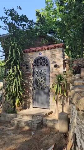 A video shows a path to a stone house with a unique door. The path is made of dirt and stones. There are trees and green plants surrounding the house. A Nazar Boncuk (evil eye charm) is visible above the door. A concrete pot with a plant is next to the door. The video pans right, revealing a path leading uphill into a wooded area with more stacked wood piles. The sun is shining, casting shadows.