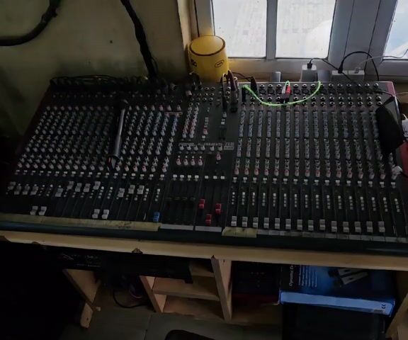 The video shows a sound mixing console and other audio equipment in a room.  The console, a large analog mixer with numerous sliders and knobs, sits on a wooden table, with a microphone resting on its surface. A yellow container is next to it. Behind the mixer, a window with a partial view outside is visible. To the left of the mixer is a stack of audio equipment, including a wireless microphone receiver, an equalizer, and possibly other amplifiers or processors. Cables connect the various pieces of equipment. The room appears to be a small studio or control room, and seems a bit cluttered. The lighting is a bit dim. There are no people visible in the video.