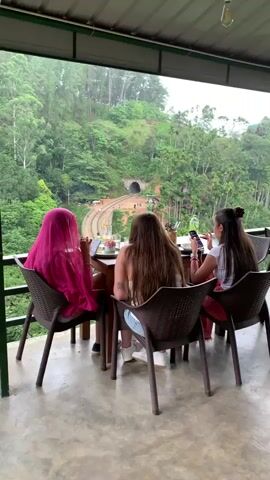 Three women are seated at a table on a covered patio, facing away from the camera. The woman on the left wears a bright pink headscarf. In front of them, the patio opens up to a view of a lush green, hilly landscape. A railway track curves through the scene, leading to a tunnel carved into the hillside. Several people are visible walking along the track and on the ground surrounding it. The women appear to be looking at their phones or engaged in conversation. Glasses and food items are on the table. The lighting suggests it might be overcast or late in the day.