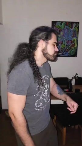 The video shows three people dancing in a room. 

The first person is a man with long dark hair and a beard, wearing a grey t-shirt with a vintage print and dark pants. He is standing in front of a white wall with a colorful abstract painting and some furniture.

The second person is a woman with long dark hair, wearing a white crop top and denim shorts. She is holding a fan and dancing in front of a white wall with several small art pieces, including ones that feature text. There is a couch behind her, and a disco light is casting colorful patterns.

The third person is a man with long dark hair, wearing a black tank top. He is dancing and holding a cell phone in front of a white wall with posters and a mirror. There are speakers visible in the background.