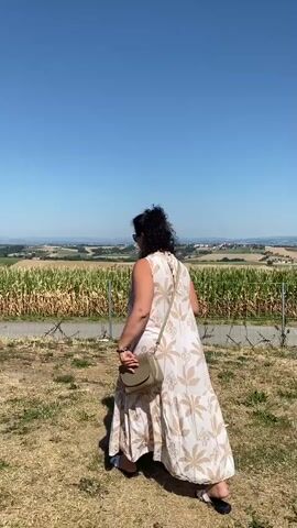 A woman with dark, curly hair is standing in a grassy field, facing away from the camera. She is wearing a long, sleeveless, light-colored dress with a floral pattern and a small, olive-green crossbody bag. She holds a dark-colored smartphone in her right hand, which she raises to take a picture. In the background, there is a clear blue sky, rolling hills, and a cornfield bordered by a wire fence. The woman then turns to face the camera, holding the phone in her left hand, and smiles.