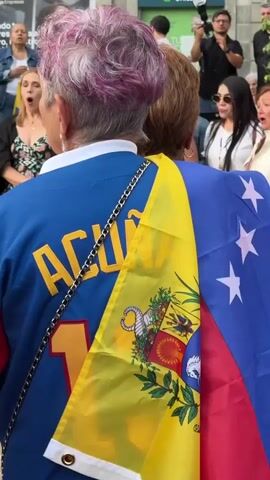 The video shows a group of people standing outdoors, possibly on a sidewalk or plaza, in front of a building with signs for "Unicaja". The focus is on the back of a person wearing a blue jacket with yellow and red lettering that says "AGUILA 10". A Venezuelan flag is draped over their shoulder. In front of this person, a group of about ten people are gathered, some of whom are singing or speaking with open mouths. One person in the group is playing a ukulele. The people in the front row are diverse in age and gender. The environment appears to be urban, with a building and signage visible in the background. The lighting suggests it is daytime, possibly overcast. The overall scene suggests a gathering or protest, perhaps with a musical element.