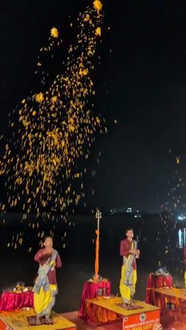 The video shows a nighttime outdoor ceremony with a dark sky and a body of water in the background. Four young men, dressed in maroon shirts and yellow lower garments, stand on elevated platforms draped in red and orange fabric. They are participating in a ritual, as indicated by the flowers being tossed into the air and the objects they hold. The men are positioned at intervals along a waterfront, each with a trident-like object on a pole behind them and small offerings on their platforms. The scene is lit by artificial lights, and the overall atmosphere suggests a religious or cultural event, possibly a form of Aarti.