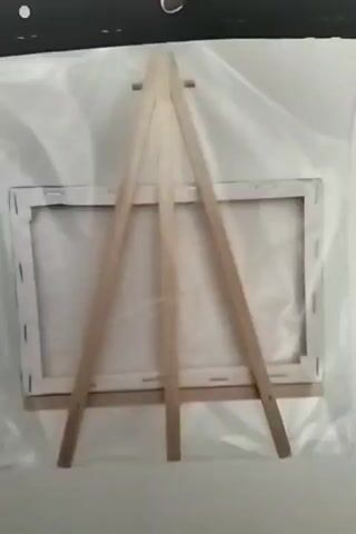 The video shows a person's hand holding up a small canvas painting of a European town with buildings reflected in a body of water. The painting is propped up on a wooden easel. The video begins with the back of the painting and easel. The camera then pans to the side, revealing the front of the painting as the hand moves the canvas. The video ends with a clear view of the painting on the easel against a white background.
