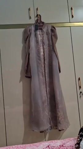 A long, beige-brown dress hangs from a dark brown plastic hanger in front of a light-colored wardrobe. The dress features an inner solid-colored, long-sleeved garment with puffed shoulders. Overlaid is a sheer, translucent outer layer adorned with intricate floral or paisley patterns in a darker brown or golden hue. A small golden brooch is pinned to the upper left side of the sheer overlay. A tag is visible near the collar. The wardrobe behind the dress has simple wooden handles. Below the dress, part of a bed with a pink and white patterned sheet is visible. The scene is indoors, likely a bedroom in Pie, Indonesia.