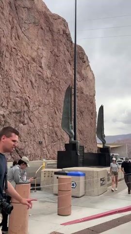 The video pans across a scenic overlook with a massive, reddish rock face on the left. In the mid-ground, a large, dark base supports two tall, metallic sculptures resembling stylized wings or hands reaching upwards, with a flagpole rising behind them. Several people are walking along a paved pathway in front of the sculptures, some interacting with each other, while others are facing away from the camera. A recycling bin is visible near the pathway. Further in the background, a road curves to the right, with cars driving on it. Several white buildings with columns and balconies are visible on the right side of the road, some adorned with large American flags. The sky is overcast with dramatic clouds. The camera movement suggests a slow, sweeping motion, capturing the scale of the landscape and the architectural elements.