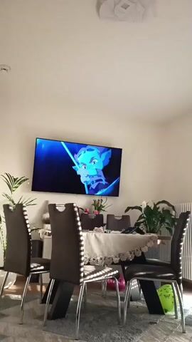The video shows a dining area with a table surrounded by chairs. A TV is mounted on the wall, displaying an animated scene with blue and teal colors. There are plants on either side of the table. Two balloons are visible on the floor near the table. The room appears to be well-lit. There are no people visible in the scene. The animation on the TV changes scenes throughout the video.