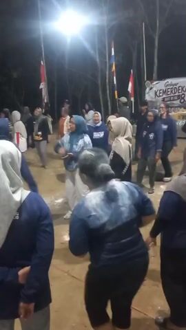 A group of people are gathered outdoors at night.  The lighting appears to be from artificial sources, possibly spotlights, illuminating the ground and the participants.  Several individuals are visible in the foreground and mid-ground, most wearing dark blue tops, some with headscarves.  One woman in the center is wearing a blue and white patterned top and light-colored pants, and she is waving.  Another person in the immediate foreground is facing away from the camera, wearing a dark blue top with white markings.  A banner is visible in the background with text that appears to include "KEMERDEKAAN" and the number "81".  There are flags and trees in the background. The ground appears to be dirt or sand. The atmosphere seems to be festive, with people engaged in some kind of activity or celebration.