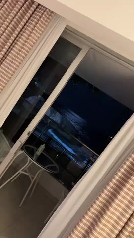 The video captures a serene nighttime view from a balcony, showcasing the ocean with gentle waves. The balcony is furnished with a small round glass table and two white chairs, with a green bottle resting on the table. Striped curtains in brown and white frame the scene, enhancing the tranquil ambiance.