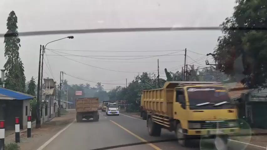 A dashcam video shows a road with several dump trucks. The video starts with the camera positioned inside a vehicle, looking out at the road ahead. Several dump trucks are visible, some in the distance and others closer to the camera. The road is paved and has yellow lane markings. On either side of the road, there are trees, utility poles with overhead wires, and some buildings. The sky is overcast and hazy. The trucks appear to be driving in the same direction. The camera vehicle is also moving. The video is grainy and the lighting is dim, suggesting it may be early morning or late evening.