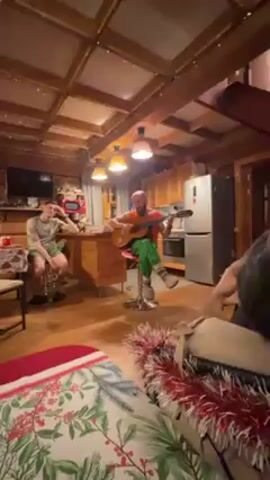 A man with a beard and a bald head is sitting on a red stool and playing an acoustic guitar in a rustic, wood-paneled kitchen area. He is wearing an orange tank top, green and white striped pants, and patterned socks with furry slippers. Two other people are present in the room. One is seated on a stool at the kitchen island, listening with his hand to his head. The other person is partially visible on a couch in the foreground, facing the man playing the guitar. The room is decorated with holiday decor, including tinsel on the couch. The kitchen includes wood cabinets, a stainless steel refrigerator, and a stove. The lighting is provided by overhead pendant lamps. The camera angle is from a low perspective, capturing the scene from the couch.
