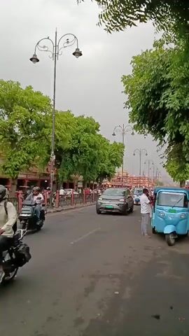 Here's a description of the video:

The video depicts a street scene in India. It's daytime, with overcast skies suggesting it might be a cloudy day. The street is lined with green trees and ornate streetlights with multiple lamp heads. On the left side of the road, there are buildings with a red facade. People are visible on the sidewalks. 

Various vehicles move along the street: cars, motorcycles, and auto-rickshaws (tuk-tuks). Some people are standing on the side of the road, including a man in a white shirt near a light blue auto-rickshaw. 

The video seems to be taken from a stationary position on the side of the road.
