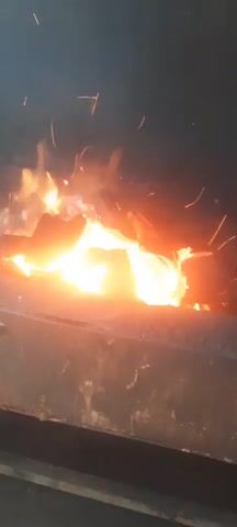 A close-up shot shows a fire burning in a dark, metal container. Sparks fly from the flames. A yellow hairdryer is held in front of the fire, its nozzle directed towards the flames. The hairdryer is being held by a person whose hand is visible. The flames are bright orange and yellow, and they flicker and dance. The container appears to be made of weathered metal. The background is dark and indistinct. The video is a short loop, repeating the same few seconds of footage.