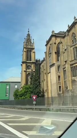 The video shows a car driving on a highway with a church on the side of the road. The church has a tall steeple and a clock. Next to the church is a building with colorful panels. The highway has multiple lanes and other cars are also driving on it. The sky is blue with some clouds.
