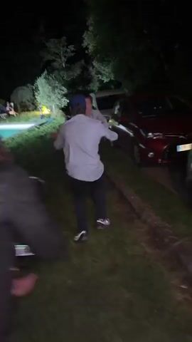 The video depicts a night scene in what appears to be a backyard with a pool. Two men are grappling with each other near the pool's edge, and they both fall to the ground. Another person, possibly filming, moves closer to the scene. In the background, there are some people gathered, seemingly watching the incident. The area is lit by artificial lights, including those illuminating the pool. There is also a vehicle parked nearby.
